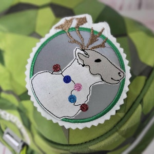 Könnte beinhalten: Ein weißes Rentier mit braunen Geweih und einer Lichterkette um den Hals. Das Rentier ist auf einem weißen Kreis mit grünem Rand und einem weißen gewellten Rand gestickt. Der Kreis befindet sich auf einem grün-weißem Tarnmuster.