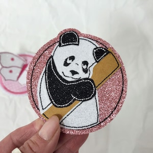Könnte beinhalten: Ein runder, glitzernder, rosa Panda-Bären-Aufnäher mit schwarzer Umrandung. Der Panda ist schwarz und weiß und hält einen braunen Bambusstiel. Ein rosa Schmetterlingsaufnäher ist teilweise im Hintergrund zu sehen.