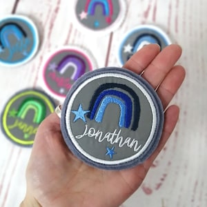 Könnte beinhalten: Ein grauer Filz-Patch mit einem blauen und schwarzen Regenbogen-Design und zwei weißen Sternen. Der Text "Jonathan" ist in Weiß unter dem Regenbogen gestickt.