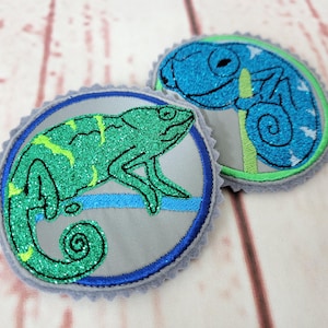 Könnte beinhalten: Zwei gestickte Aufnäher mit grünen und blauen Glitzer-Chamäleons auf grauem Hintergrund. Die Chamäleons sitzen auf einem Ast mit blauen und grünen Akzenten.