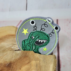 Könnte beinhalten: Ein grüner Außerirdischer mit drei Augen und einem zahnigen Grinsen ist auf einem grauen Filzkreis mit einem gelben Stern gestickt. Der Außerirdische zeigt mit einer Hand ein Peace-Zeichen und hat eine grüne Glitzer-Oberfläche.