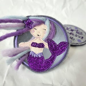 Könnte beinhalten: Ein runder Stickaufnäher mit einer Meerjungfrau mit lila glitzerndem Schwanz und Bikini-Oberteil, helllila und blauem Garnhaar sowie einem kleinen lila Sternakzent. Ein kleinerer ovaler Aufnäher mit dem Text "marie" ist daneben zu sehen.