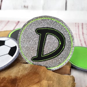 Könnte beinhalten: Ein runder, silberner Glitzeraufnäher mit dem schwarzen Buchstaben "D" und neongrünen Nähten. Der Aufnäher hat einen neongrünen Rand. Weitere Aufnäher, darunter ein Fußball-Design, sind im Hintergrund zu sehen.