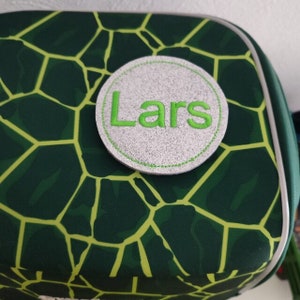 Könnte beinhalten: Eine grün-gelb gemusterte Tasche mit einem kreisförmigen Aufnäher. Der Aufnäher ist grau mit dem Namen "Lars" in Grün gestickt. Die Tasche hat ein geometrisches Design.