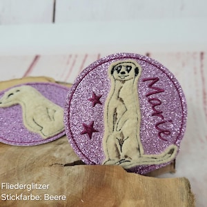 Könnte beinhalten: Ein pinkfarbener Glitzer-Aufbügel-Patch mit einem weißen gestickten Erdmännchen, zwei roten Sternen und dem Namen "Marie" in weißer Stickerei.