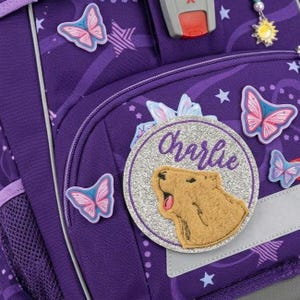 Könnte beinhalten: Ein violetter Rucksack mit Sternen- und Wirbelmuster, verziert mit mehreren rosa Schmetterlingsaufnähern, einem Sonnenanhänger und einem runden silbernen Glitzeraufnäher mit dem Namen Charlie und einer Capybara-Illustration.