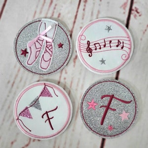 Könnte beinhalten: Vier runde, silberne Glitzer-Aufbügelpatches mit rosa Nähten. Ein Patch zeigt ein Paar rosa Ballettschuhe mit einem rosa Band. Ein weiterer Patch zeigt eine Musiknote mit einem Violinschlüssel. Ein dritter Patch zeigt eine rosa Girlande mit dem Buchstaben F. Der letzte Patch zeigt den Buchstaben F mit rosa Sternen.