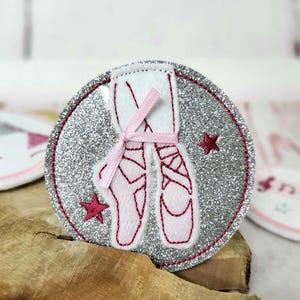 Könnte beinhalten: Ein runder, glitzernder Aufnäher mit einem Paar rosa Ballettschuhen, die mit rosa Bändern gebunden sind. Das Design ist von einem rosa Rand umgeben und enthält zwei rosa Sternakzente.