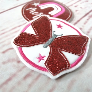 Könnte beinhalten: Rote Glitzer-Schmetterlings-Applikation mit weißen und rosa Akzenten. Der Schmetterling hat einen weißen Körper und schwarze Fühler. Die Applikation ist von einem rosa Kreis mit weißen Sternen umgeben.
