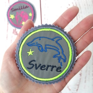 Könnte beinhalten: Grauer Filz-Kreis mit einem grün und blau gestickten Delfin, einem gelben Stern und dem Namen "Sverre" in Schwarz.