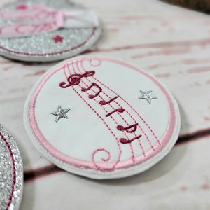Könnte beinhalten: Ein runder weißer Patch mit rosa Nähten am Rand. Der Patch hat ein rosa Notenbild mit einem Violinschlüssel und Sternen.