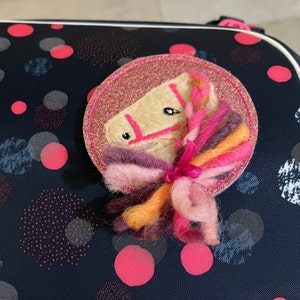 Könnte beinhalten: Ein rosa und weißer Filz-Pferdekopf auf einem rosa Glitzer-Hintergrund. Das Pferd hat eine rosa Mähne und einen rosa Schwanz aus Garn.