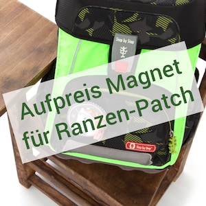 Könnte beinhalten: Ein grün-schwarz-gelber Rucksack mit einem roten Knopf und dem Schriftzug "Step by Step" auf der Vorderseite. Der Rucksack hat einen Magneten und einen Patch mit der Aufschrift "Aufpreis Magnet für Ranzen-Patch".