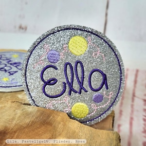 Könnte beinhalten: Ein runder, silberner Glitzer-Aufnäher, bestickt mit dem Namen "Ella" in lila Schrift. Der Aufnäher hat gelbe und lila gestickte Punkte und rosa florale Akzente. Geeignet für Kleidung oder Accessoires.