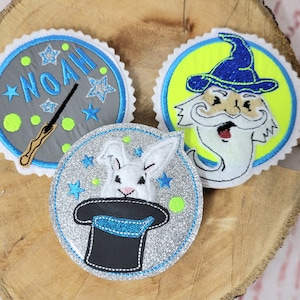 Könnte beinhalten: Drei gestickte Bügelbilder. Ein Patch ist grau mit blauen Sternen und dem Namen "NOAH" in blau. Ein weiterer Patch ist silber mit blauen Sternen und einem weißen Kaninchen, das aus einem schwarzen Zylinderhut schaut. Der dritte Patch ist gelb mit einem blauen Stern und einem weißen Zauberer mit einem blauen Hut und einem langen weißen Bart.