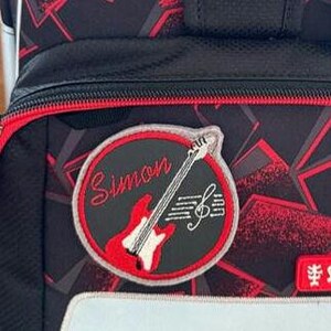 Könnte beinhalten: Nahaufnahme eines schwarz-roten Rucksacks mit einem Gitarren- und Noten-Patch. Der Patch trägt den Namen "Simon" in Schreibschrift. Der Rucksack hat ein geometrisches Muster und rote Verzierungen.