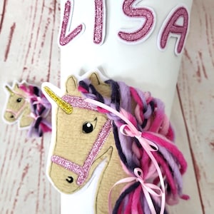 Könnte beinhalten: Ein weißer, kegelförmiger Gegenstand mit dem Namen "LISA" in rosa Glitzerbuchstaben. Ein hellbrauner Einhornkopf mit einem rosa Glitzerzaumzeug und einem goldenen Horn ist befestigt. Das Einhorn hat eine Mähne aus rosa, lila und dunkelblauem Garn, die mit rosa Bändern gebunden ist.