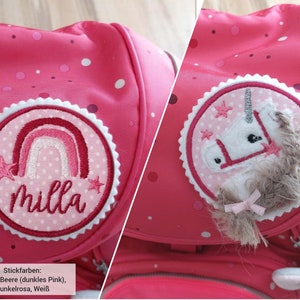 Könnte beinhalten: Ein leuchtend pinker Rucksack mit zwei runden Stickaufnähern. Ein Aufnäher zeigt einen Regenbogen und den Namen Milla in Schreibschrift. Der andere zeigt einen weißen Einhornkopf mit strukturierter Mähne und kleinen rosa Sternen auf Punkten.
