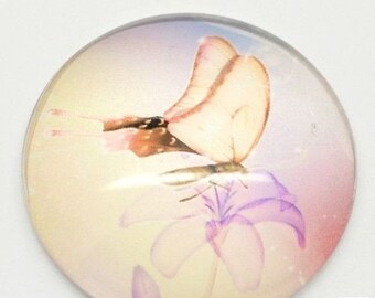 Puede incluir: Un cabujón redondo de vidrio con un fondo rosa, morado y amarillo que presenta una mariposa con marcas marrones y negras posada sobre una flor.