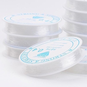 Op de afbeelding: Een pakket van acht spoelen met doorzichtige elastische draad. Elke spoel is gelabeld met "Strong & Stretchy 0.4mm Crystal Line 100%".