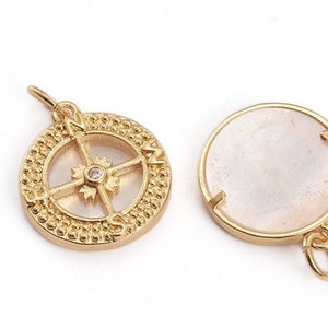 Könnte beinhalten: Goldener Kompass-Anhänger mit einem klaren Kristall in der Mitte und einer weißen Perlmuttrückseite. Der Kompass-Anhänger hat einen perlenbesetzten Rand und die Buchstaben N, S, E und W sind um den Kompass herum sichtbar.
