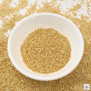 May include: A white bowl filled with gold seed beads. The beads are small and round, and they are scattered around the bowl.