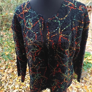 Könnte beinhalten: Schwarze Strickjacke mit offenem Frontdesign und farbenfroher Kettenstichstickerei in Rot, Orange und Grün. Der langärmlige Pullover wird auf einer schwarzen Schaufensterpuppe präsentiert und zeigt den strukturierten Strickstoff und das aufwendige Design.