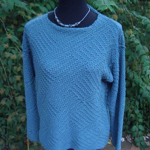 Könnte beinhalten: Türkisfarbener Strickpullover mit langen Ärmeln, präsentiert auf einer schwarzen Schaufensterpuppe. Der Pullover hat ein strukturiertes Muster und einen Rundhalsausschnitt. Eine Perlenkette mit grünen und silbernen Perlen liegt um den Hals der Schaufensterpuppe.