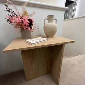 May include: A light wood side table with a white book and a vase with dried pink and burgundy flowers. The vase is a light beige ceramic with two handles.