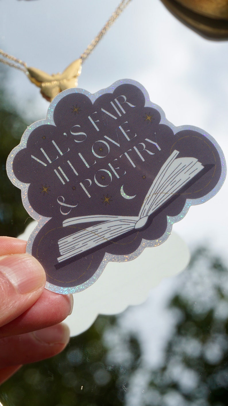 May include: A holographic sticker with a dark purple background featuring a white open book with the text "All's Fair in Love & Poetry" in white with gold stars and a crescent moon.