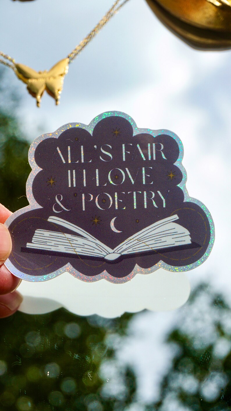 May include: A holographic sticker with a cloud shape and a book with a crescent moon. The sticker says "All's fair in love & poetry".