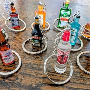 May include: A collection of miniature liquor and beer bottle keychains featuring various brand labels including Budweiser, Jack Daniel's, Baileys, Jägermeister, Bombay Sapphire, Smirnoff, Grand Marnier, and Fireball, each attached to a silver metal ring.