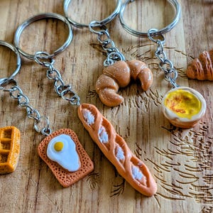 May include: A selection of miniature food keyrings, featuring a waffle, toast with a fried egg, a baguette, a croissant, and a fried egg in a small dish. The keyrings are attached to silver key rings and chains.