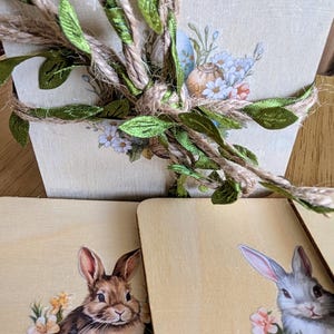May include: Four wooden coasters with a light brown background. Each coaster features a different image of a rabbit with flowers. The coasters are tied together with twine and green leaves.