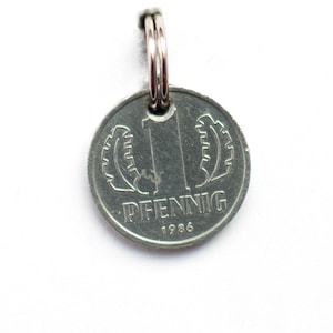 Può includere: Una moneta d'argento da 1 Pfennig del 1986 con un portachiavi. La moneta presenta un grande numero "1" circondato da una corona di foglie.
