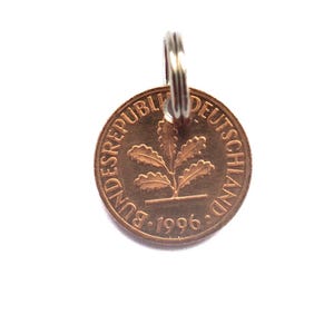 Op de afbeelding: Een koperkleurige ronde munthanger met een afbeelding van een eikentakje in het midden. De tekst BUNDESREPUBLIK DEUTSCHLAND en het jaartal 1996 staan in reliëf langs de rand. Een zilverkleurige metalen ring is aan de bovenkant bevestigd.