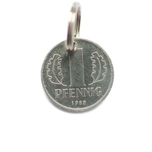 Könnte beinhalten: Eine silberne deutsche 1-Pfennig-Münze aus dem Jahr 1985 mit einem Schlüsselring. Die Münze zeigt eine große Ziffer "1" und das Wort "PFENNIG" auf der Vorderseite.