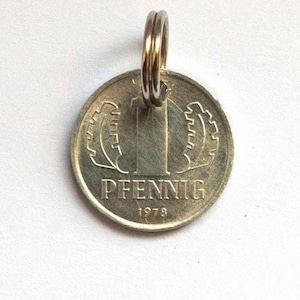Puede incluir: Un colgante de moneda de color plateado con un anillo dorado. La moneda presenta la palabra "PFENNIG" y el año "1978". La moneda tiene un diseño de dos hojas o engranajes estilizados a cada lado del número "1".