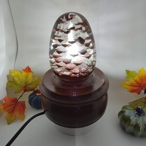 May include: A wooden table lamp with a clear glass pine cone shaped shade. The lamp is lit and the light shines through the glass. The lamp is on a wooden base and has a black cord.