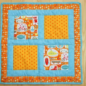May include: A blue and orange patchwork quilt with a variety of colorful designs, including a robot, a bicycle, a submarine, and a rocket. The quilt is made of different fabrics and has a quilted texture.