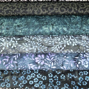 May include: A stack of fabric bolts featuring various patterns and colors. Designs include floral, abstract, and geometric shapes in shades of black, white, blue, and purple. The fabrics appear to be cotton or a similar material, suitable for quilting or crafting.