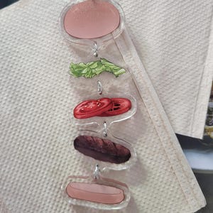 May include: A clear acrylic keychain featuring a layered burger design. The keychain includes a top bun, lettuce, tomato slices, a burger patty, and a bottom bun, all connected by small metal rings and a silver clasp.
