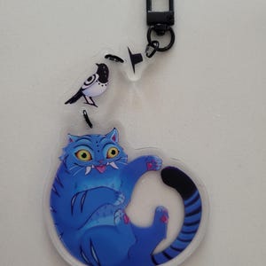 以下が含まれることがあります： 青い猫のキーホルダーと白黒の鳥のチャーム。猫は黄色い目と鋭い歯をしています。キーホルダーには黒い留め金が付いています。