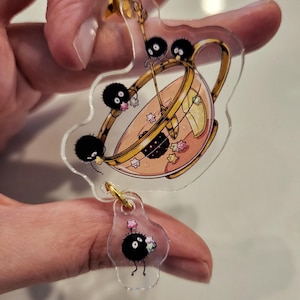 Puede incluir: Un llavero de acrílico transparente con una taza de té llena de una bebida rosa y una rodaja de limón. Cuatro duendes de hollín negros y blancos rodean la taza de té, y uno cuelga de la parte inferior del llavero.