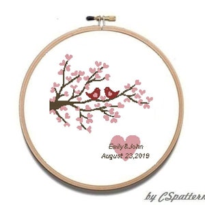 Puede incluir: Un bastidor de bordado de punto de cruz con un dise&ntilde;o de dos p&aacute;jaros rojos posados en una rama floreciente. El dise&ntilde;o incluye flores rosas en forma de coraz&oacute;n, dos corazones rosas y los nombres "Emily & John" con la fecha "23 de agosto de 2019".
