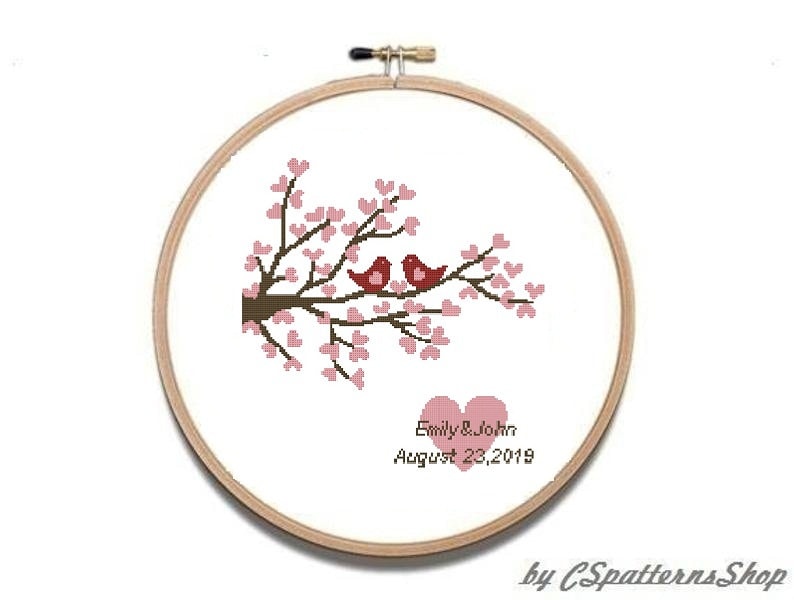 Puede incluir: Un bastidor de bordado de punto de cruz con un dise&ntilde;o de dos p&aacute;jaros rojos posados en una rama floreciente. El dise&ntilde;o incluye flores rosas en forma de coraz&oacute;n, dos corazones rosas y los nombres "Emily & John" con la fecha "23 de agosto de 2019".