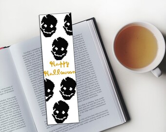 Puede incluir: Un marcapáginas blanco con un diseño pixelado en blanco y negro de calaveras y el texto "Happy Halloween" en letras doradas.