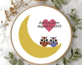 Pode incluir: Um bordado em ponto cruz num bastidor de madeira redondo com uma lua crescente amarela, um coração rosa e duas corujas. O texto diz Emily & John August 23, 2025. O design é bordado em tecido branco com figuras de corujas castanhas e roxas.
