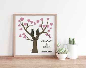 May include: A framed cross-stitch artwork featuring a brown tree with two black cat silhouettes perched on a branch. The tree has pink heart-shaped leaves. The text reads Elisabeth & Oliver and the date 20.05.2021 in black lettering.