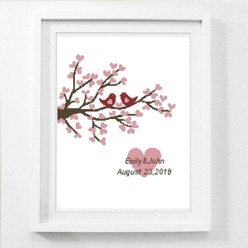 Puede incluir: Imagen enmarcada de punto de cruz que representa una rama de &aacute;rbol con flores rosas y dos p&aacute;jaros rojos. Debajo, un coraz&oacute;n rosa y los nombres "Emily & John" con la fecha "August 23, 2019" est&aacute;n bordados en negro.